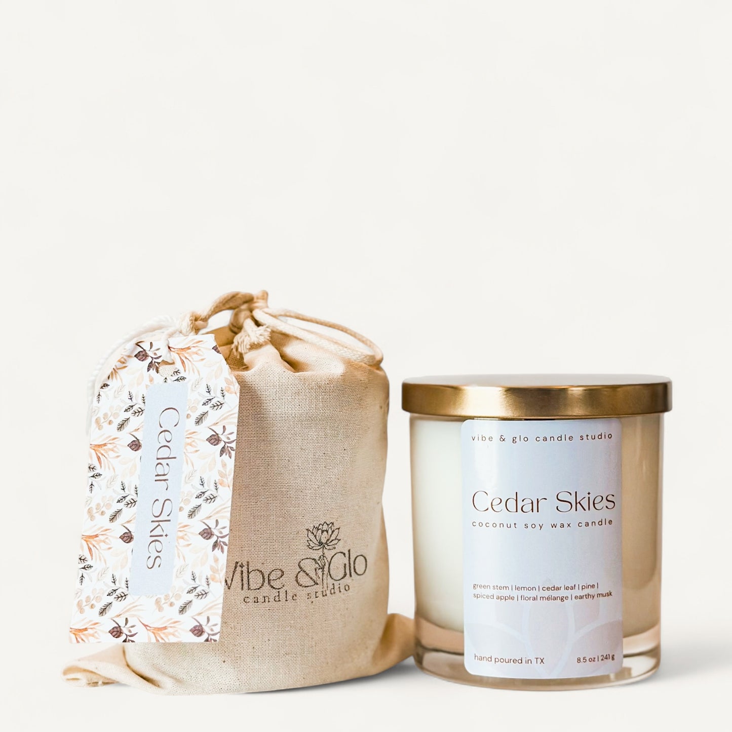 Cedar Skies candle in glass jar with gold lid, accompanied by a natural fabric pouch and tag from Vibe & Glo Candle Studio.
