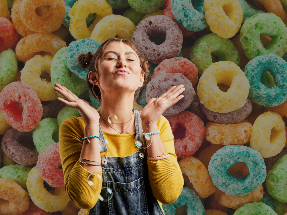Playful young woman with pigtail buns and bright expression blowing kisses in front of a colorful fruit cereal backdrop, wearing a yellow sweater and denim overalls.