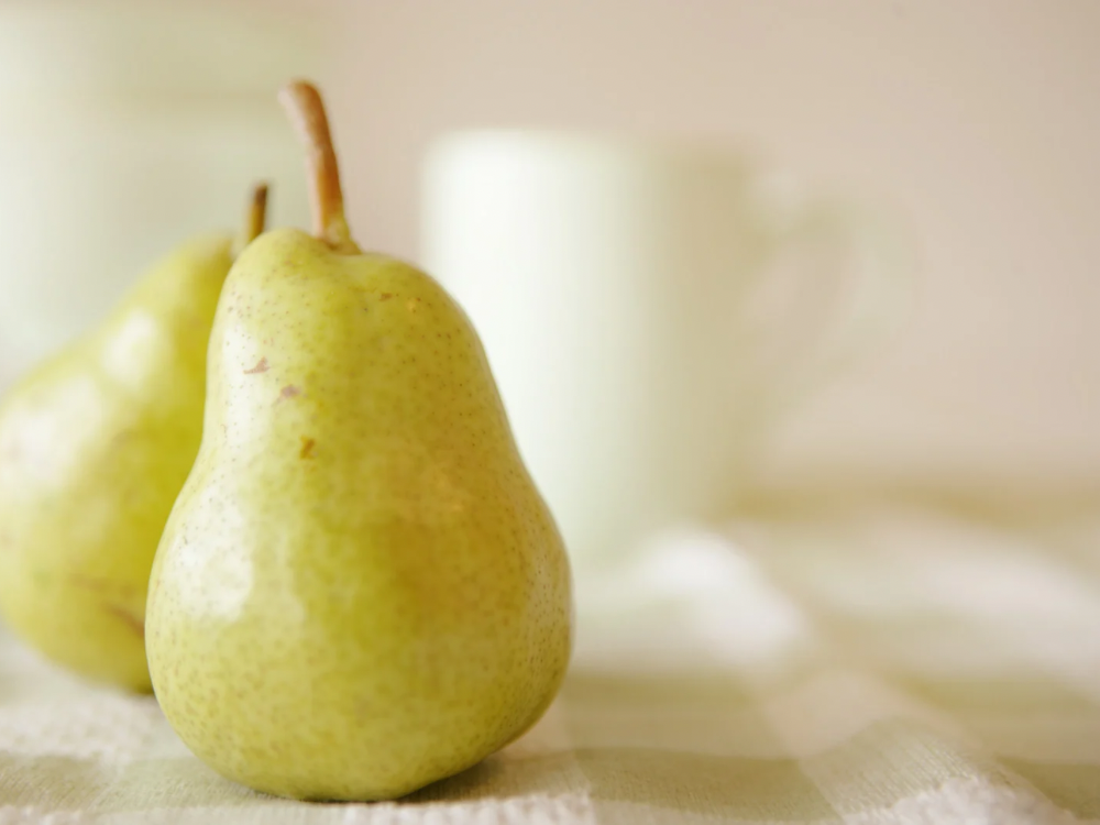 Close-up of fresh green pears on a light checkered cloth with a blurred ceramic mug in the background, creating a soft and minimal breakfast scene.