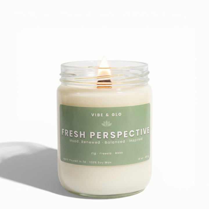Fresh Perspective 16 oz wooden wick soy candle by Vibe & Glo with fig, freesia, and moss, lit wooden wick in clear glass jar, high-key studio product photography on white background.