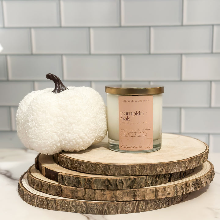 Candle labeled 'Pumpkin Oak' on a wooden stand with a white pumpkin against a tiled wall background.