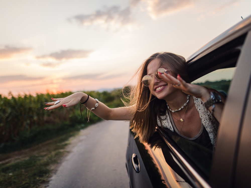 Carefree young woman smiling and enjoying the breeze with her arms out of a car window during a golden hour road trip.