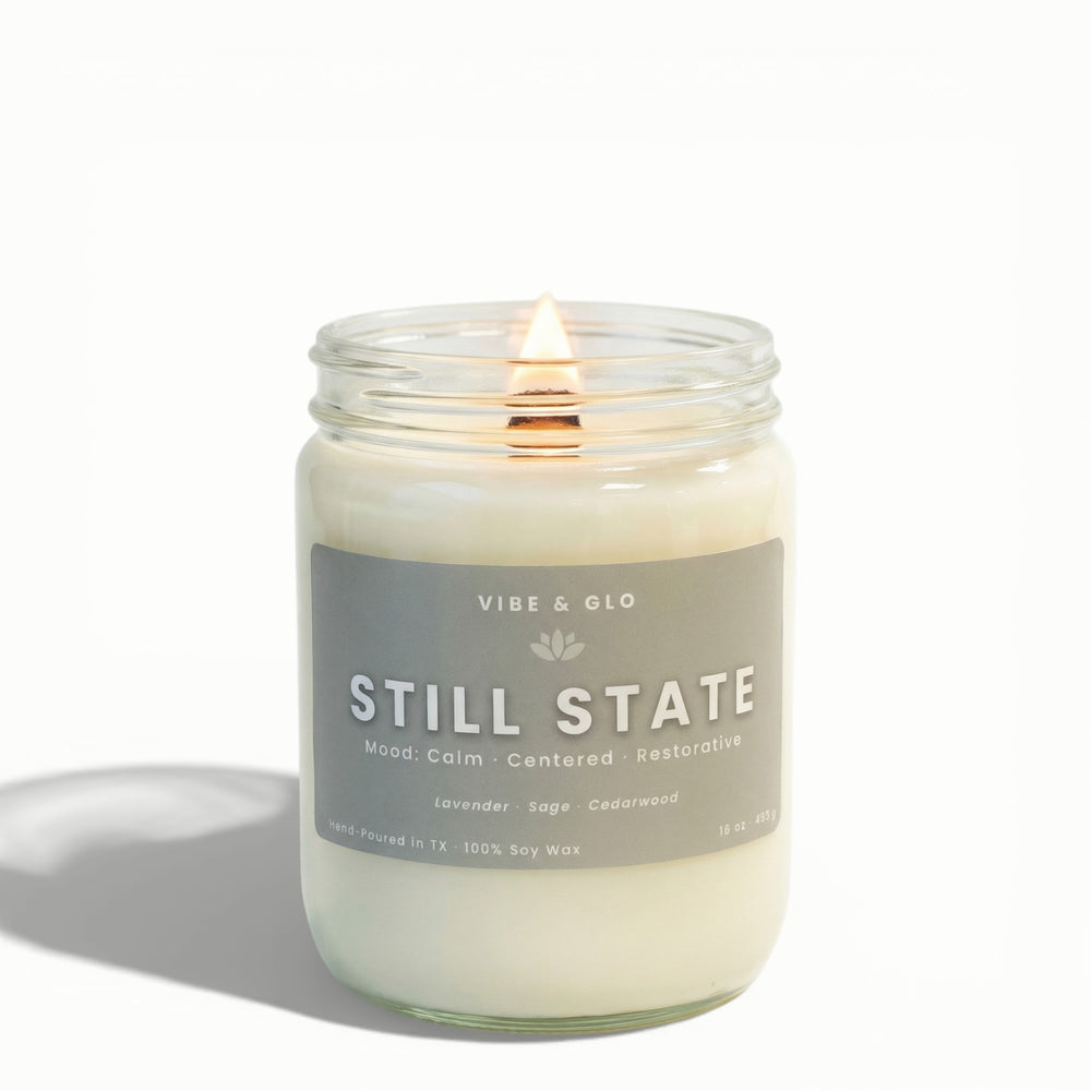 Still State 16 oz wooden wick soy candle by Vibe & Glo with lavender, sage, and cedarwood, lit wooden wick in clear glass jar, clean white background product photo.