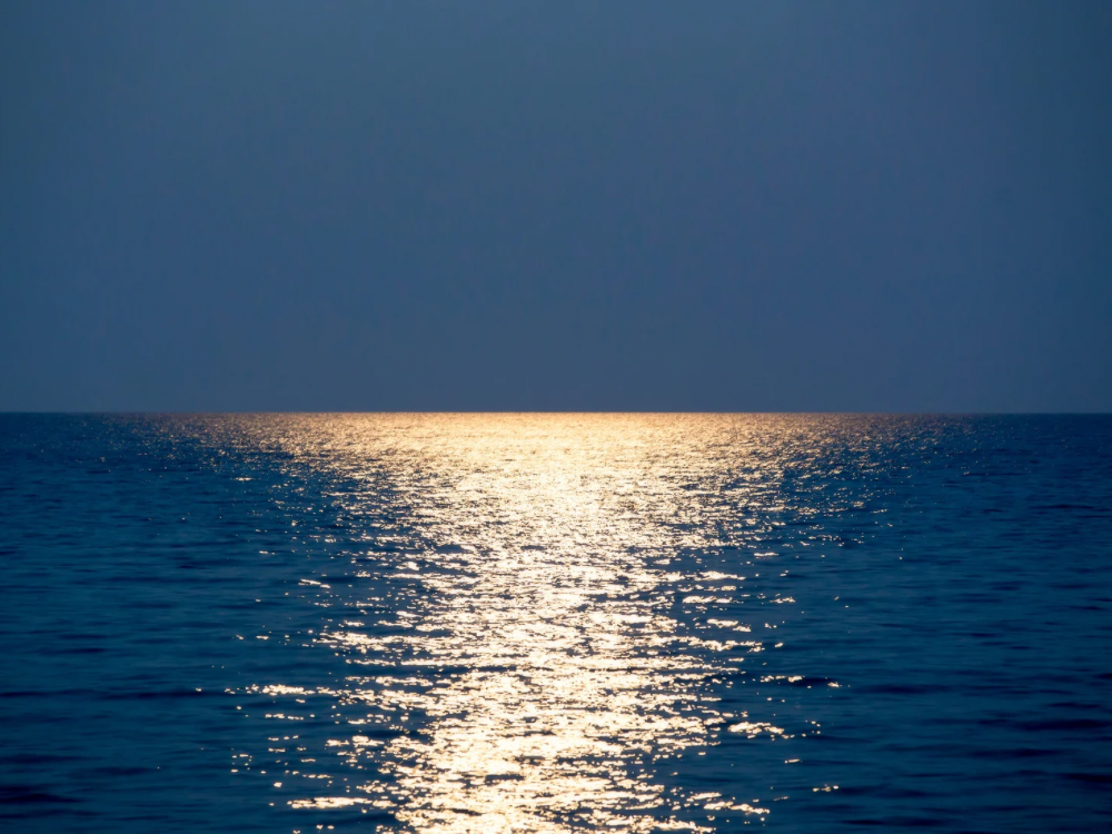 Golden sunset reflecting across a calm blue ocean, creating a peaceful and serene horizon under a cloudless sky.