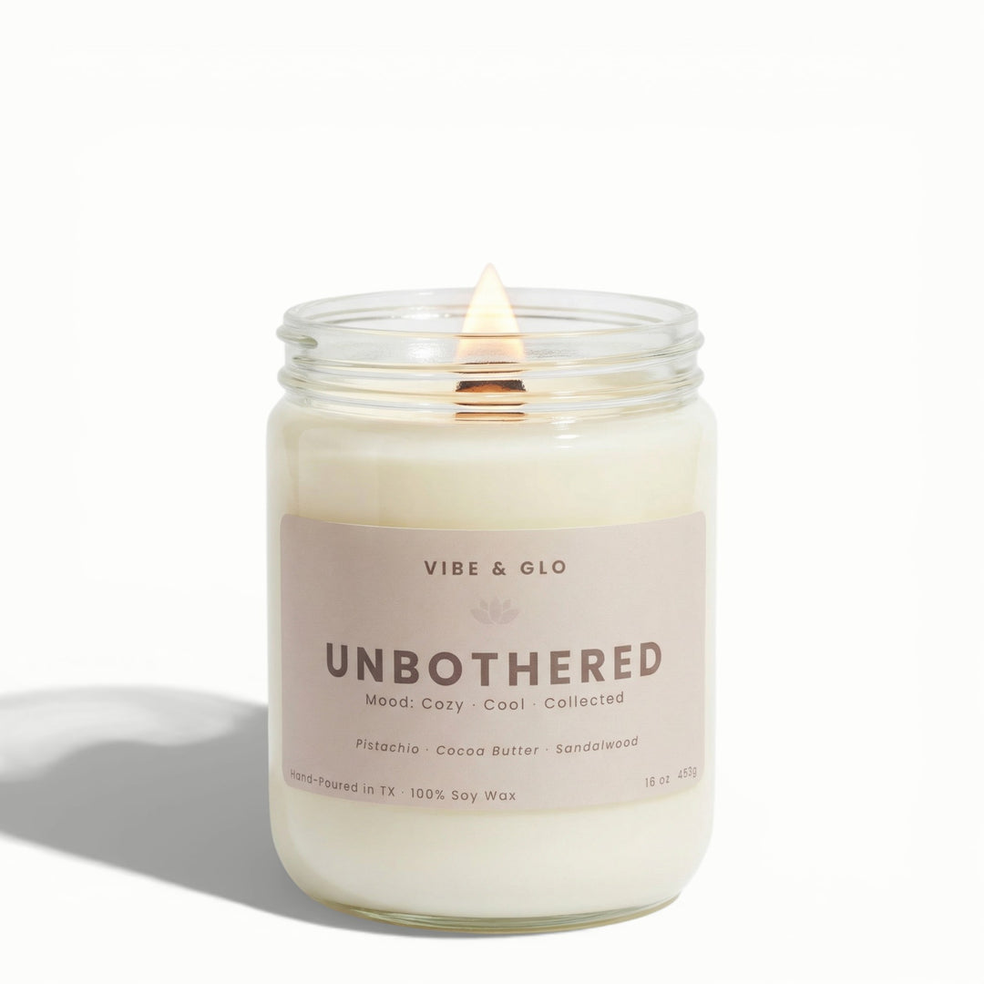 Unbothered 16 oz wooden wick soy candle by Vibe & Glo with pistachio, cocoa butter, and sandalwood, lit wooden wick in clear glass jar, centered high-key product photography.