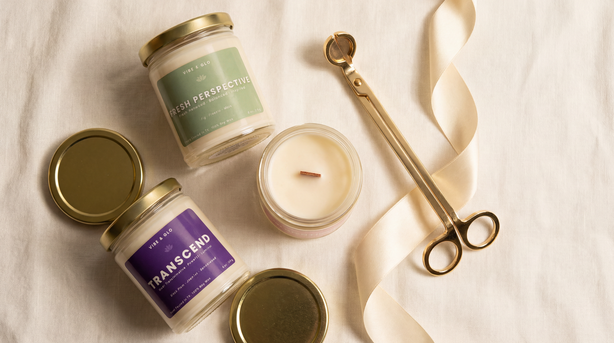 Flatlay of Vibe & Glo wooden wick soy candles including Fresh Perspective and Transcend with gold lids and wick trimmer on neutral linen background.