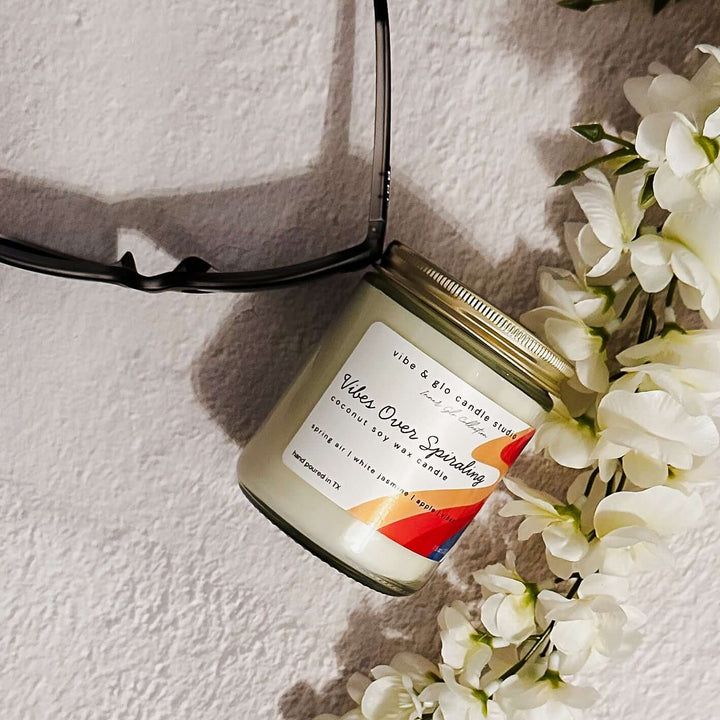 Vibes Over Spiraling coconut soy wax candle styled with sunglasses and white flowers. Fresh, floral scent with spring air, jasmine, and apple.
